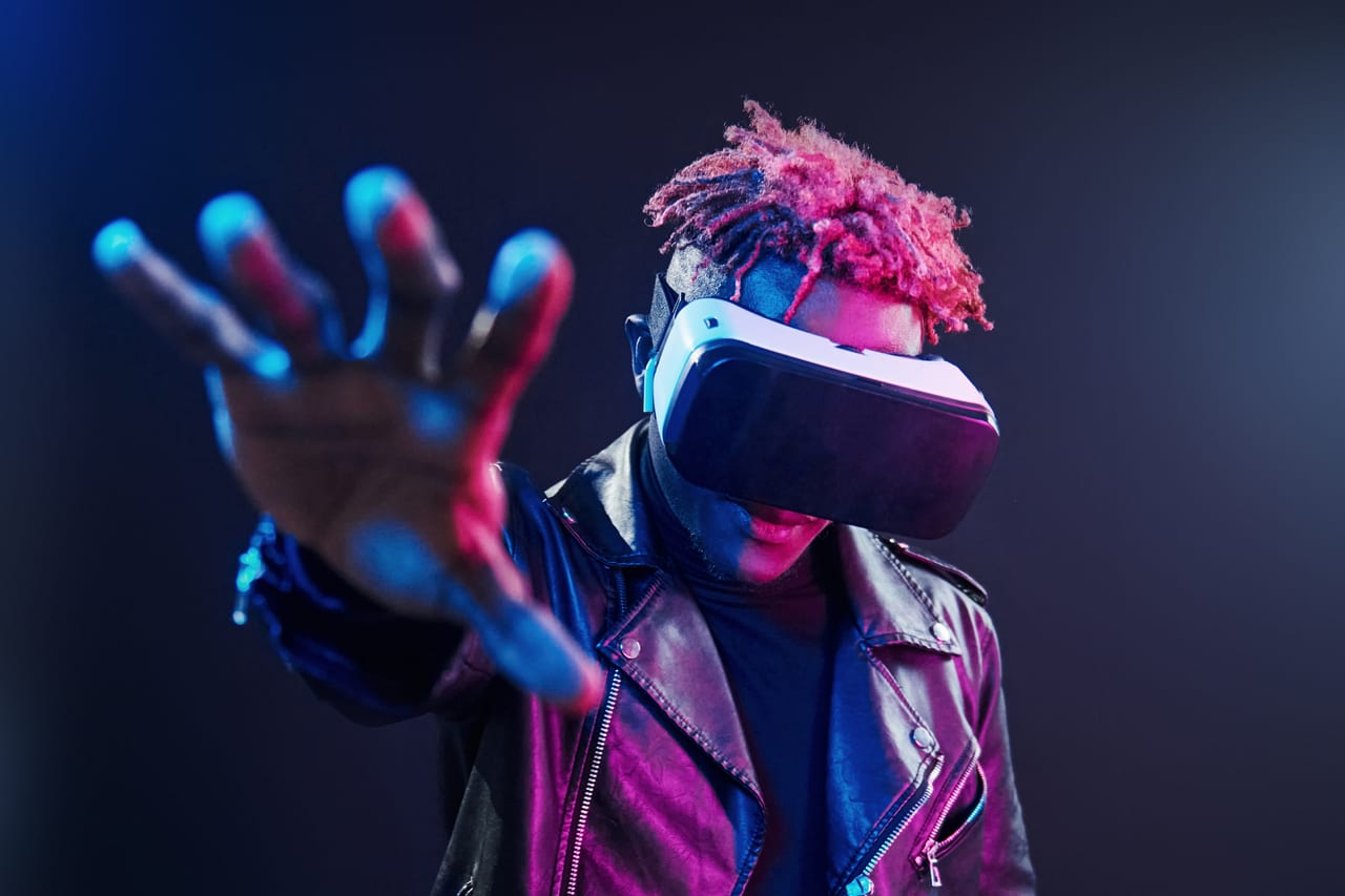 A person in a VR headset reaching a hand toward the camera in neon-lit darkness